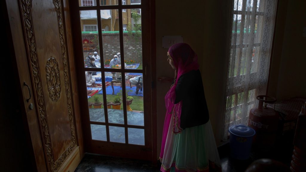 A Kashmiri girl watches from inside her house as men offer Eid prayers in the premises of a residential building in Srinagar. [Image- AP]