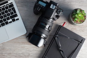 Flat lay image of a laptop, professional camera, black notebook with glasses, and a small succulent plant on a wooden desk